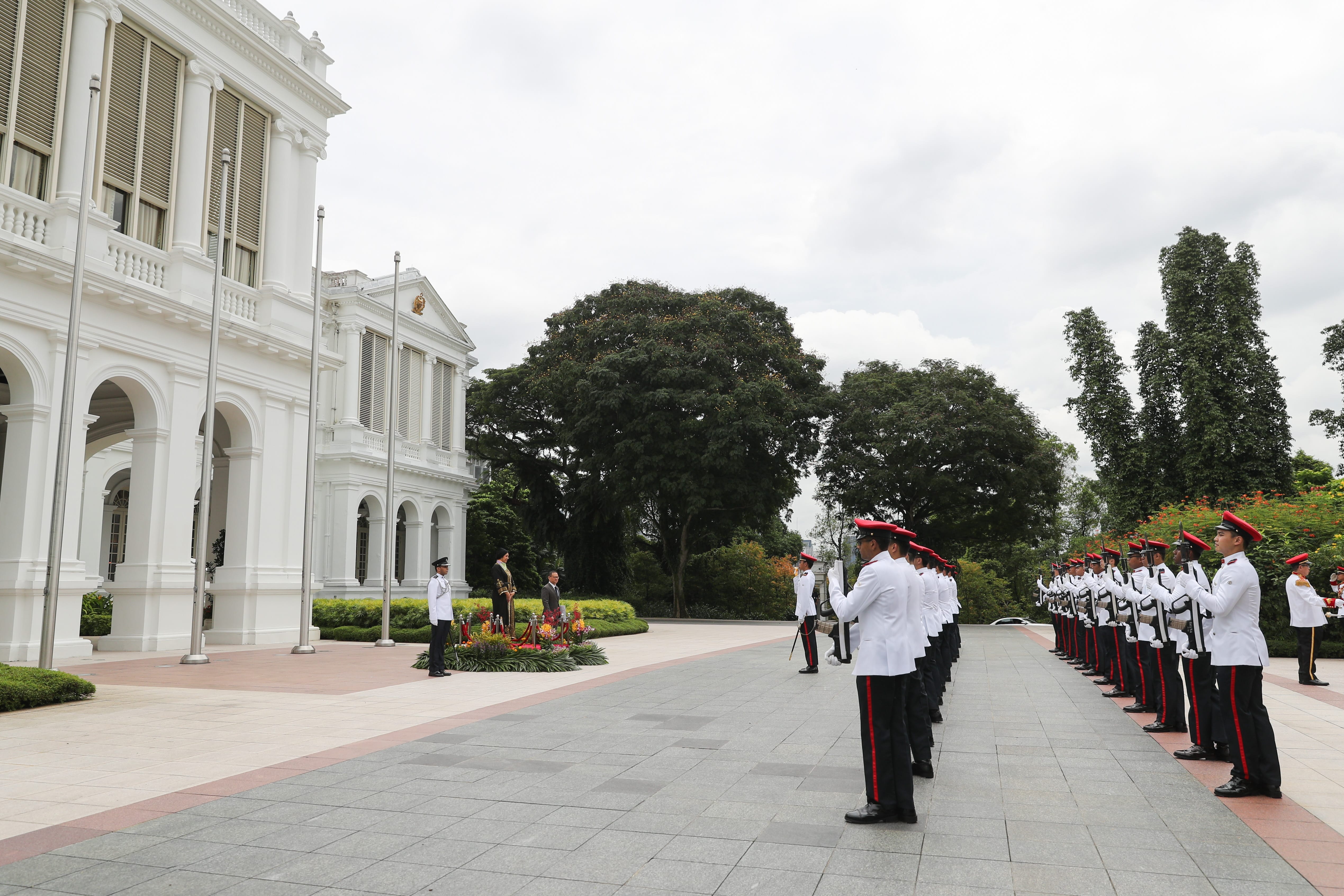 Guards in white uniforms and red hats stand at attention near a white building.
